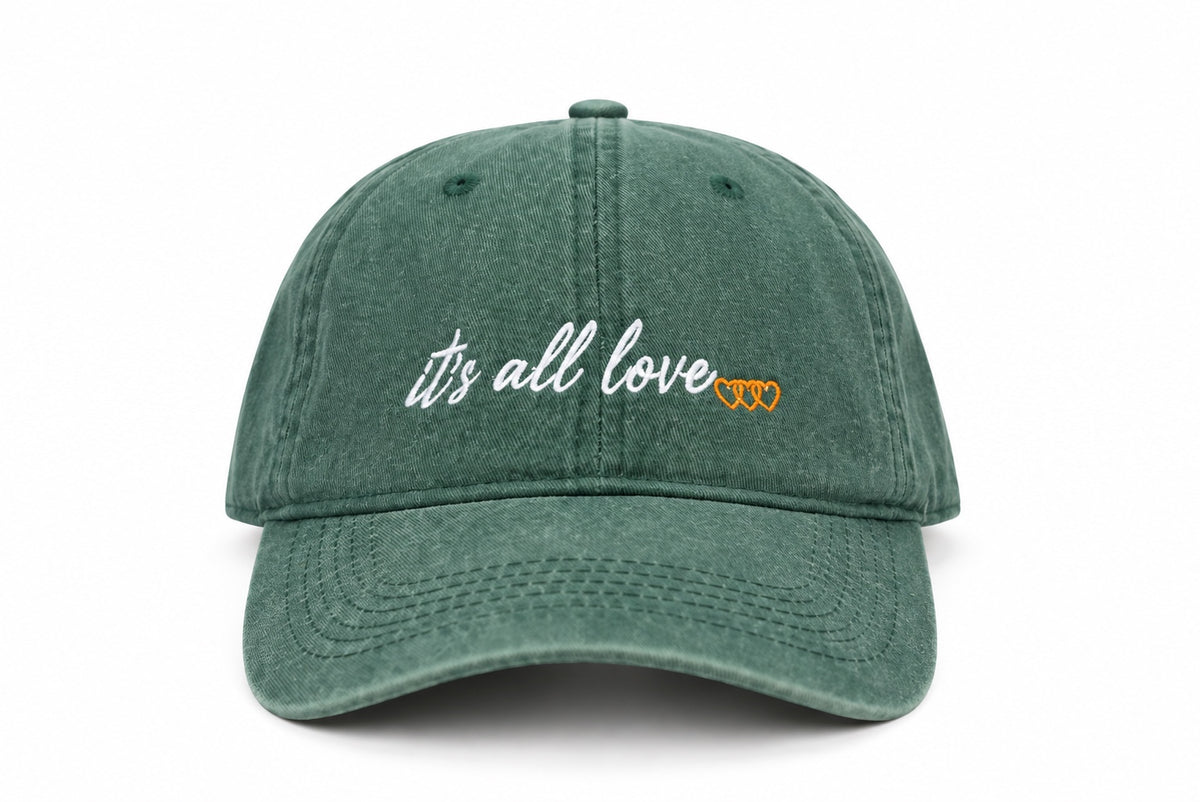 Washed green dad hat with white it’s all love script and three orange signature hearts embroidered on front