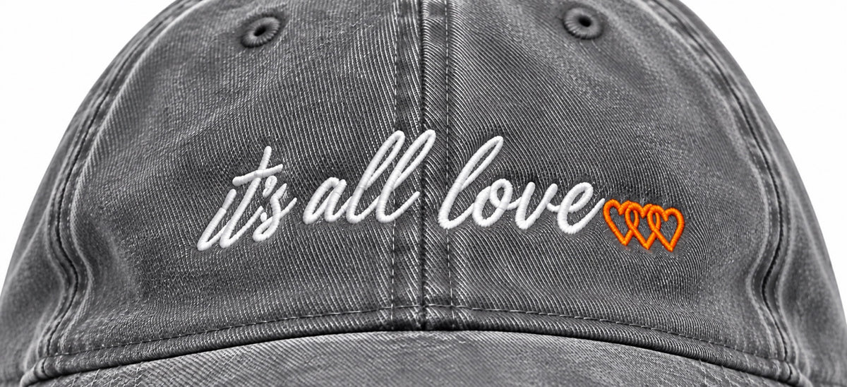 Close-up of white it’s all love script and three orange signature hearts embroidered on washed grey dad hat